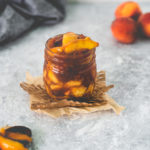 A jar of Fresh Peach Pie Filling sitting on top of a square of parchment and meshed paper with a spoonful sitting in front and a bunch of peaches behind.