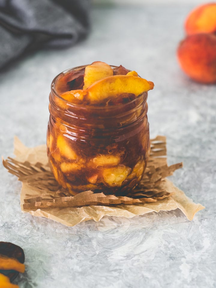 A jar of Fresh Peach Pie Filling sitting on top of a square of parchment and meshed paper with a spoonful sitting in front and a bunch of peaches behind.