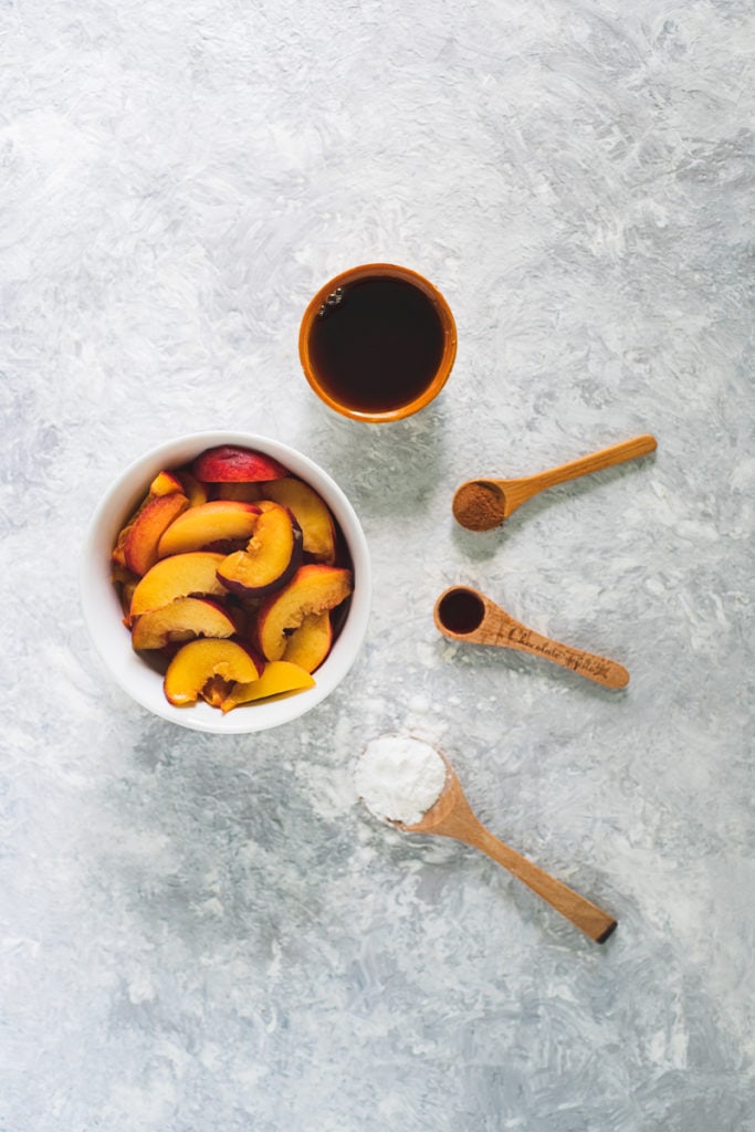 A bowl of freshly sliced peaches with a bowl of maple syrup and spoonfuls of arrowroot, vanilla extract and cinnamon surrounding in a half circle.