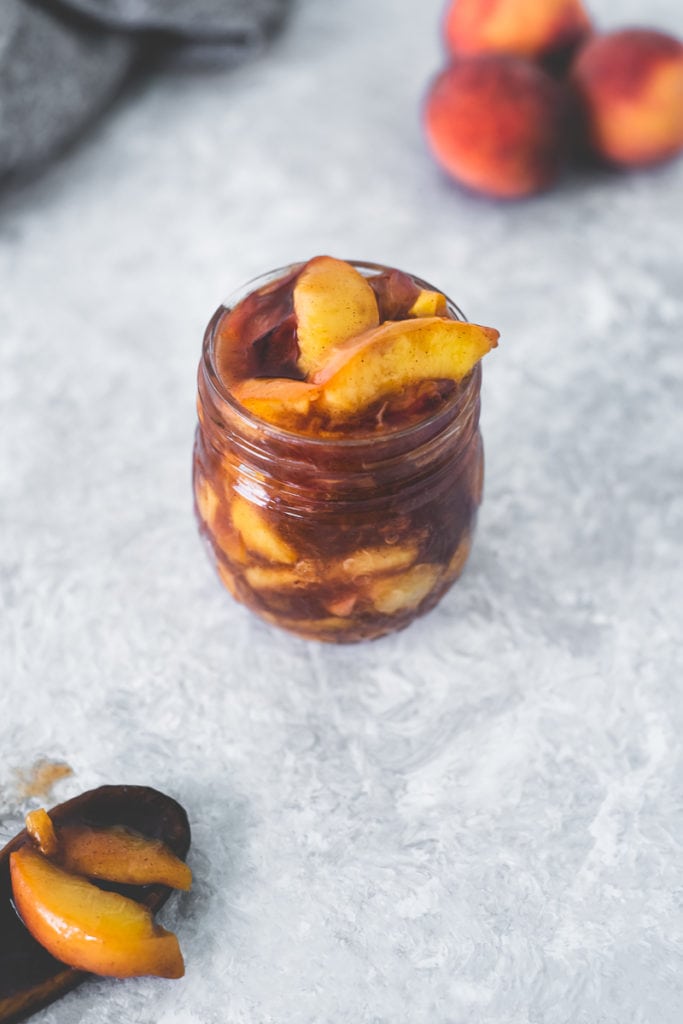 An overhead view of a heaping jar of Homemade Peach Pie Filling with a spoonful spilled next to it, along with a few whole peaches and a cloth behind.