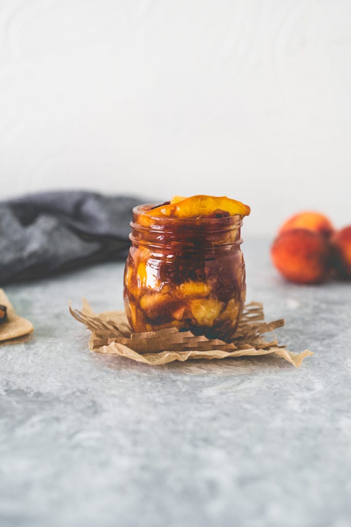 A jar filled to the top with Healthy Homemade Peach Pie Filling with a bunch of peaches, a wooden spoon and linen cloth behind.