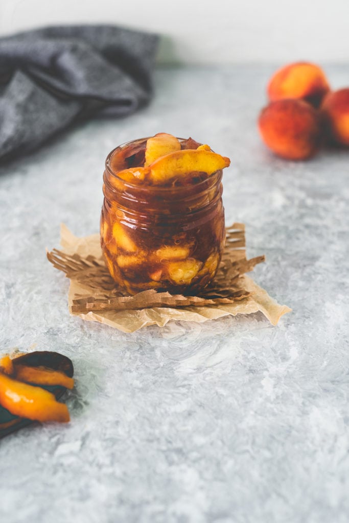 A jar of Fresh Peach Pie Filling sitting on top of a square of parchment and meshed paper with a spoonful sitting in front and a bunch of peaches behind.