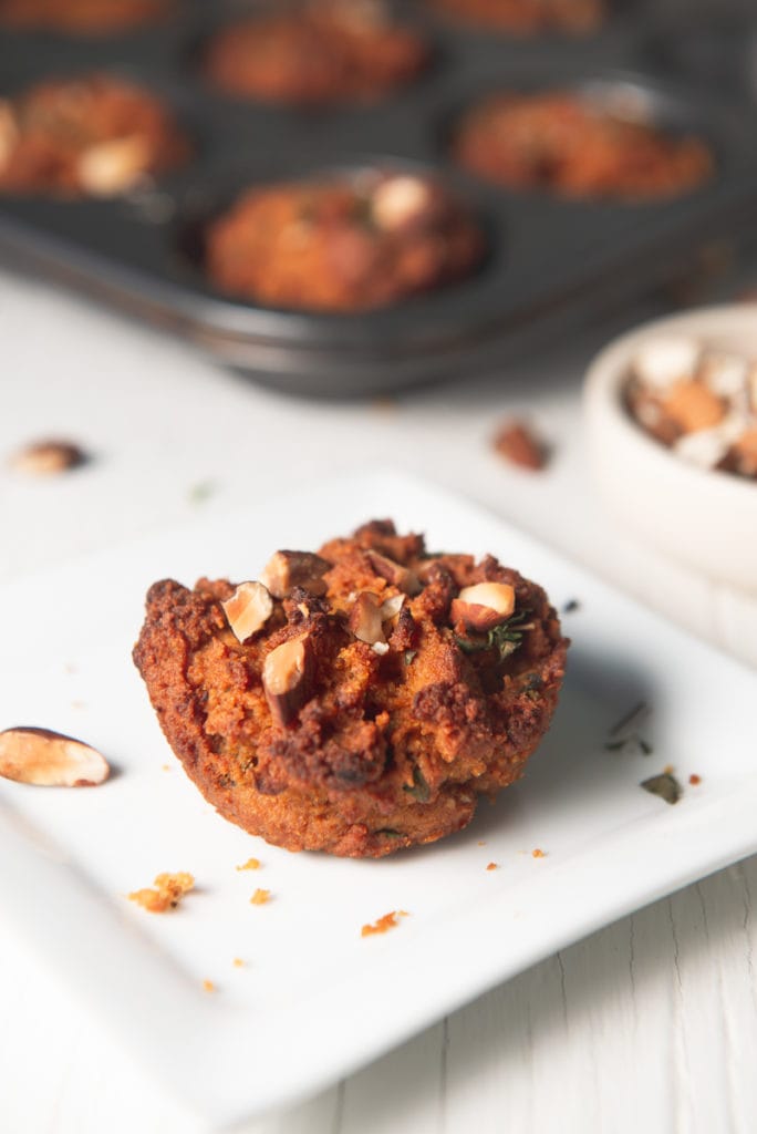 A sweet potato muffin sitting on a plate in front of a full muffin tin and a bowl of chopped almonds.