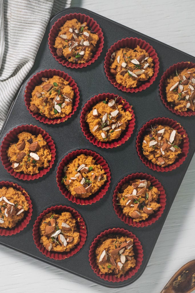A muffin tin filled with muffin batter topped with chopped almonds and fresh thyme.