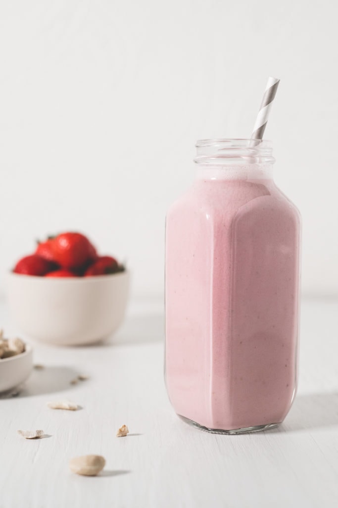 A pink smoothie in a jar with a straw sitting beside an overfilled bowl of cashews and a bowl of fresh strawberries.