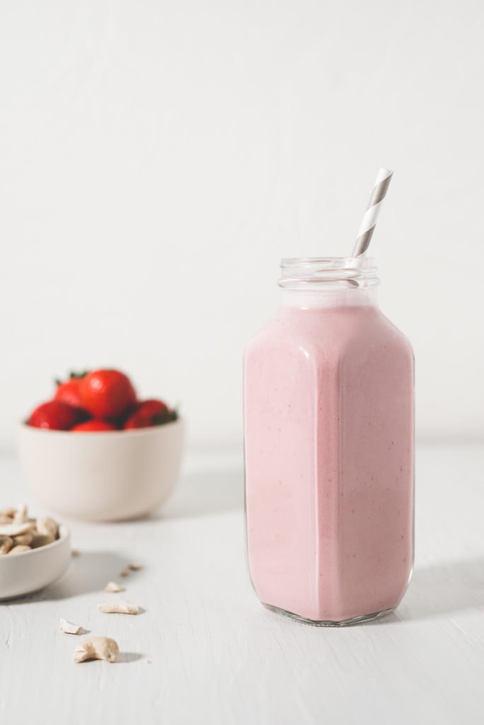 A beautiful, pink Strawberry Shortcake Smoothie, freshly poured with a straw, sitting beside raw ingredients.