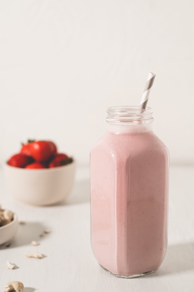 A freshly poured, strawberry smoothie sitting in front of a bowl of strawberries and spilled cashews.