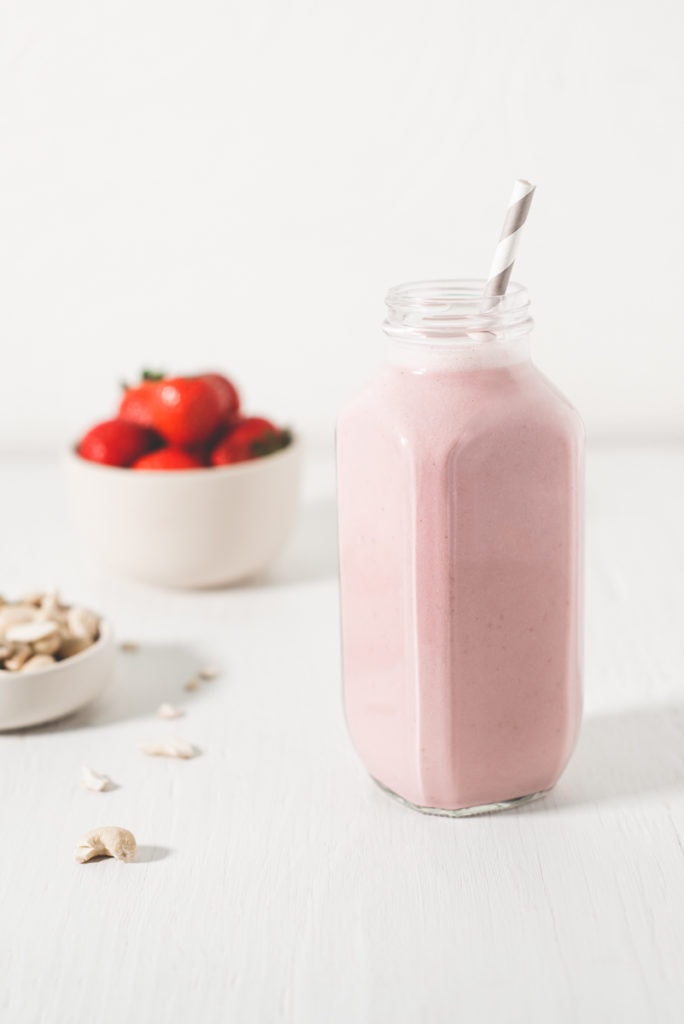 A healthy strawberry smoothie beside raw cashews and a bowl of strawberries.