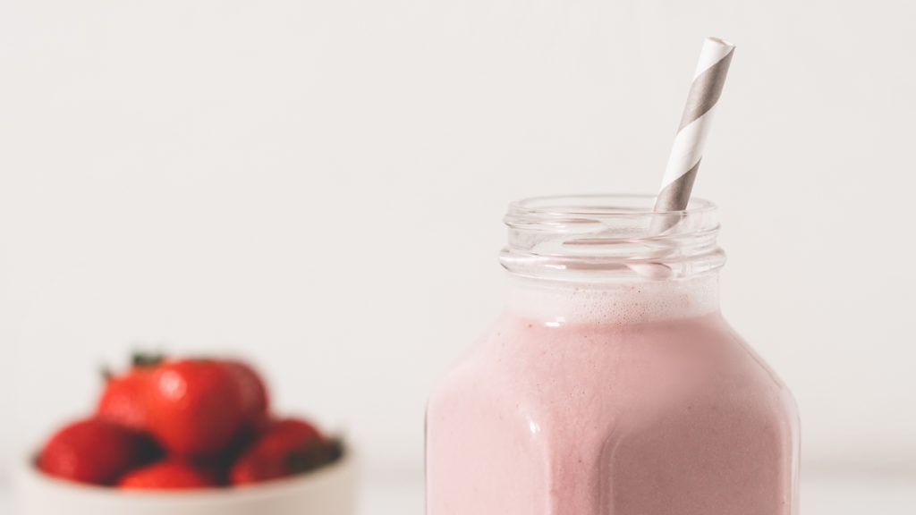 A pink strawberry smoothie in a glass bottle with a striped straw in front of a bowl of ripe strawberries.