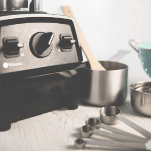 A blender, small pot, measuring cups, a colander and a wooden spoon sitting on a counter, ready to be used.