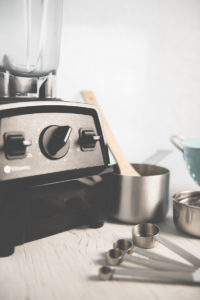 A blender, small pot, measuring cups, a colander and a wooden spoon sitting on a counter, ready to be used.