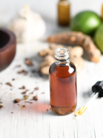 A full tincture with the dropper sitting beside along with a mortar and pestle, limes, ginger, garlic and loose herbs spilled about.