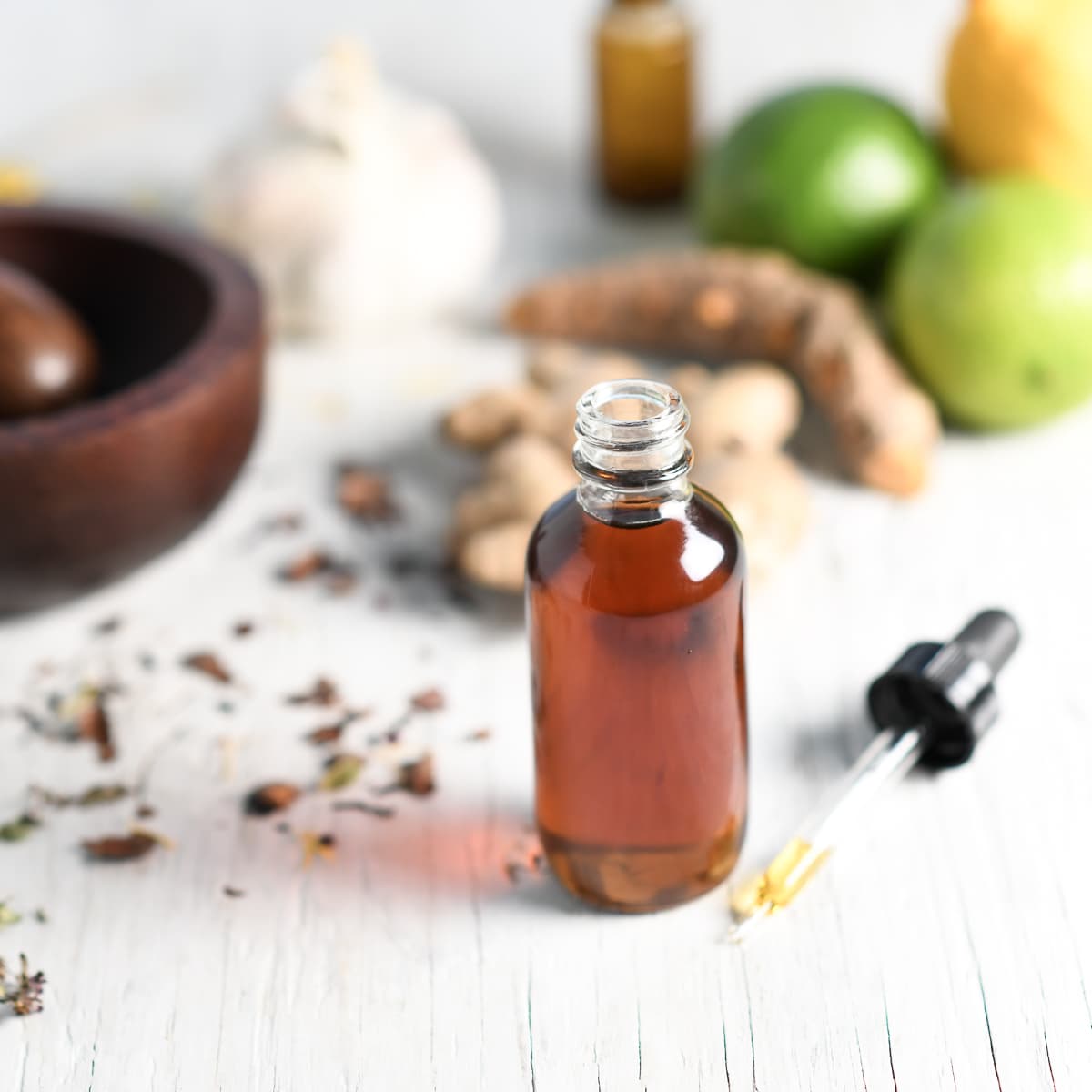 A full tincture with the dropper sitting beside along with a mortar and pestle, limes, ginger, garlic and loose herbs spilled about.