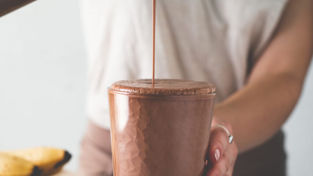 A chocolate smoothie being poured into a tall glass.