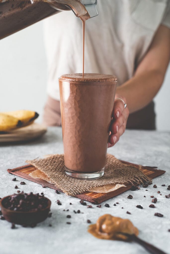 A chocolate smoothie being poured into a tall glass with bananas, cacao nibs and a spoonful of peanut butter on the table surrounding.