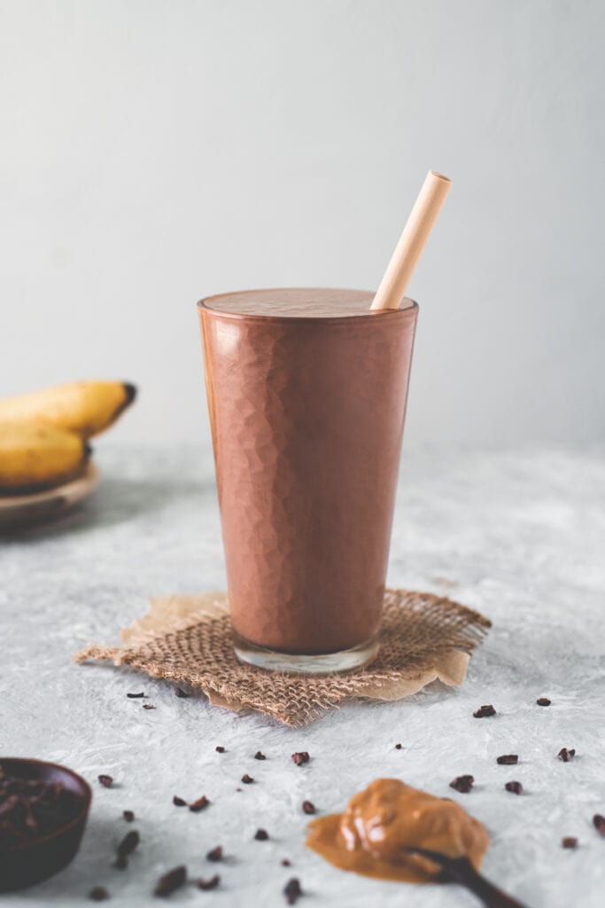 A tall glass filled to the top with a chocolate peanut butter banana smoothie sitting in the middle of a spoonful of peanut butter, a bunch of bananas and a spilled bowl of cacao nibs.