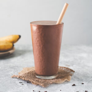 A tall glass filled to the top with a chocolate peanut butter banana smoothie sitting in the middle of a spoonful of peanut butter, a bunch of bananas and a spilled bowl of cacao nibs.