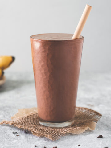 A tall glass filled to the top with a chocolate peanut butter banana smoothie sitting in the middle of a spoonful of peanut butter, a bunch of bananas and a spilled bowl of cacao nibs.