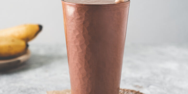 A tall glass filled to the top with a chocolate peanut butter banana smoothie sitting in the middle of a spoonful of peanut butter, a bunch of bananas and a spilled bowl of cacao nibs.