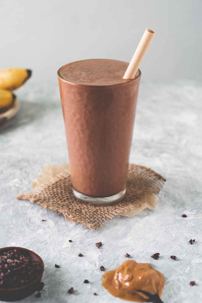 A tall glass filled to the brim with a rich chocolate smoothie and a coconut straw.