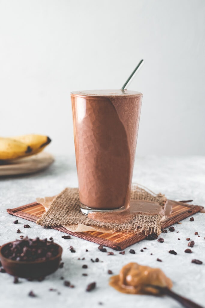 An overflowing chocolate smoothie surrounded by spilled and scattered cacao nibs, a bunch of bananas and a heaping spoonful of natural peanut butter.