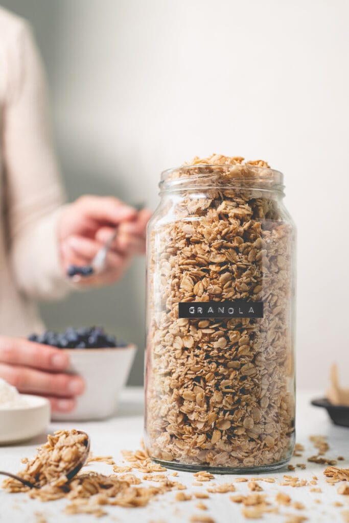 A labelled jar filled with granola that's spilling out, next to toppings and bowl of blueberries being delved into.