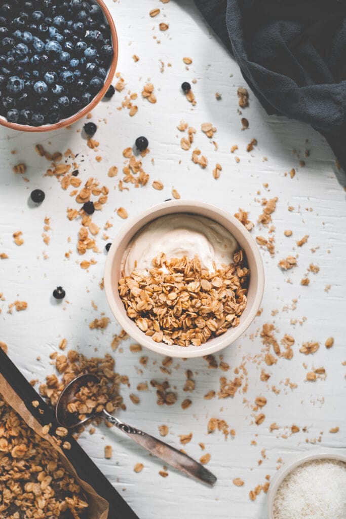 A bowl of yogurt and granola surrounded by an overflowing spoonful, a baking sheet, a bowl of blueberries, shredded coconut and a linen cloth.