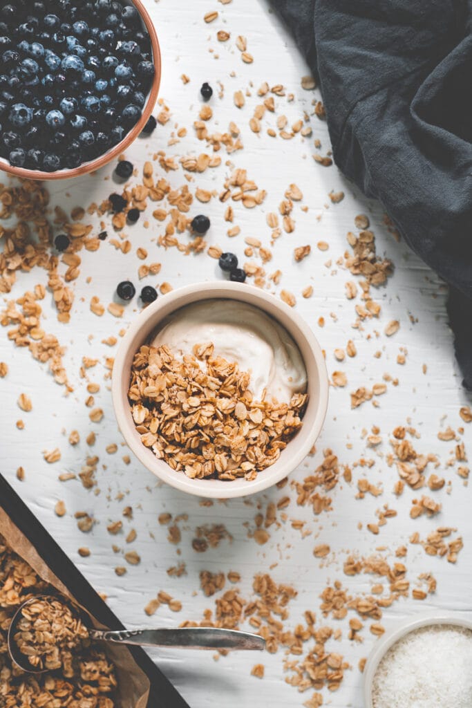 A bowl of yogurt topped with granola surrounded by spilled oats, an overflowing bowl of blueberries a linen cloth and a baking sheet with more freshly baked granola.