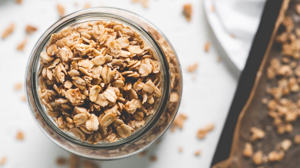 A jar overflowing with granola sitting beside a baking sheet holding more freshly made granola.