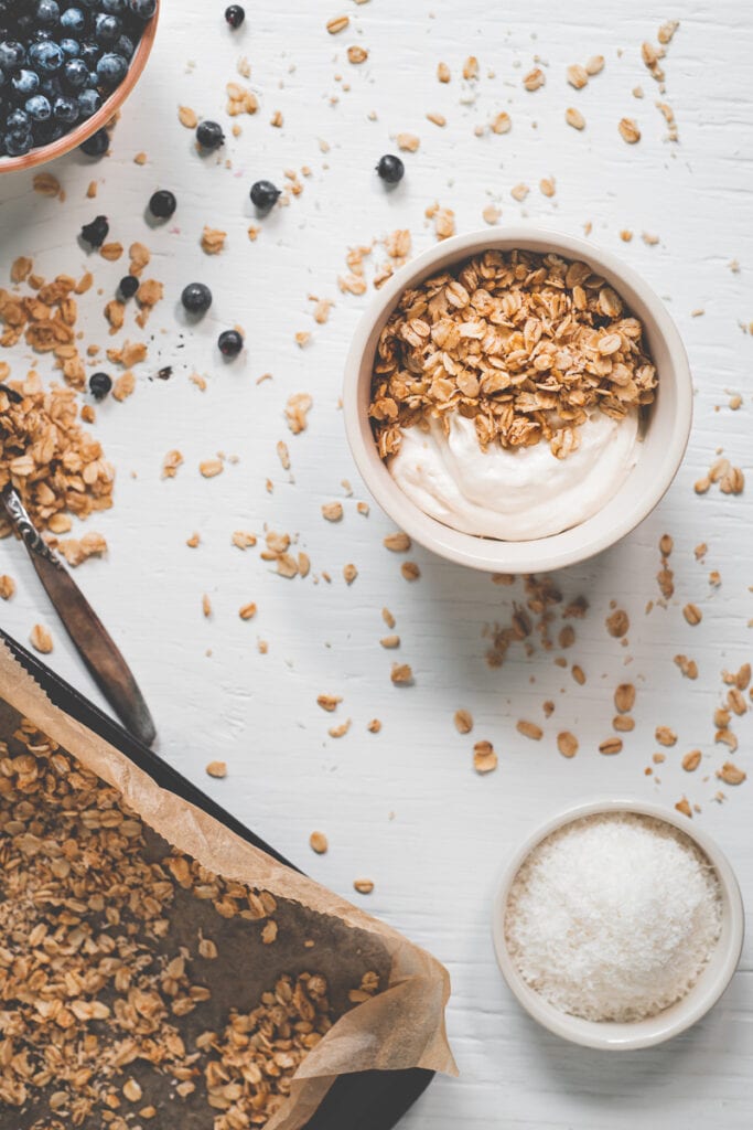 A bowl of yogurt and granola beside a bowl of coconut, a baking sheet with freshly baked granola, a bowl of overflowing blueberries and spoonful of spilled oats.