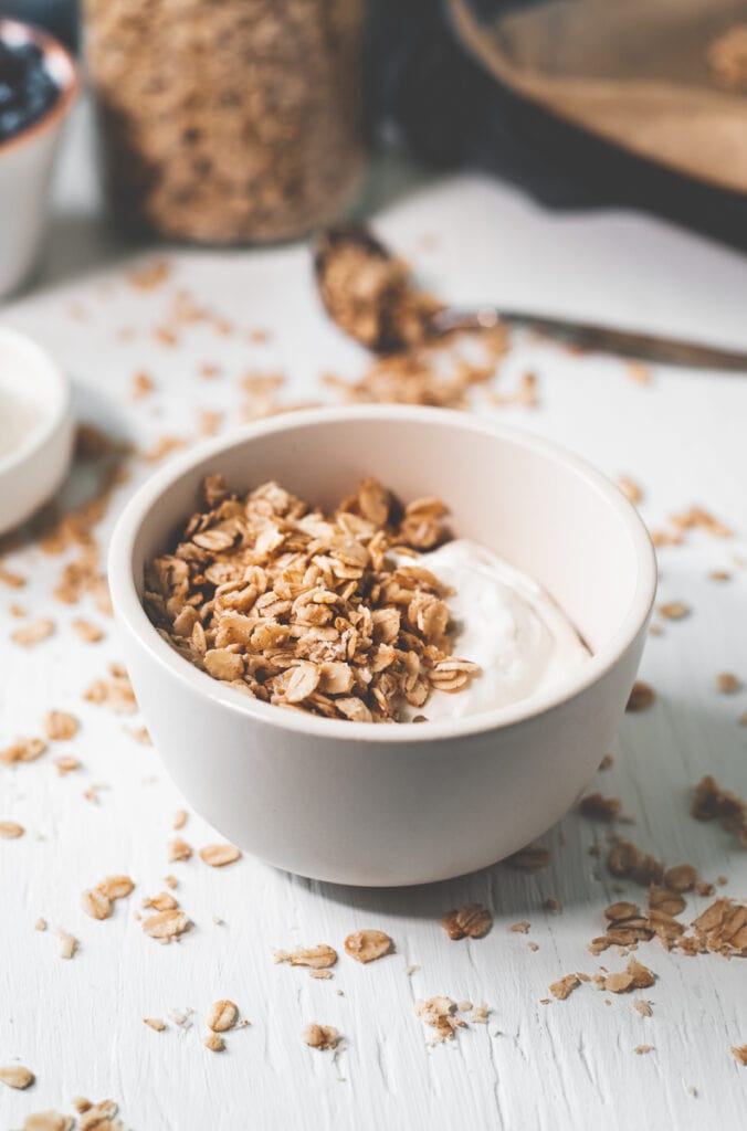 A bowl filled with yogurt topped with granola sitting in front of a spoon of spilled granola.