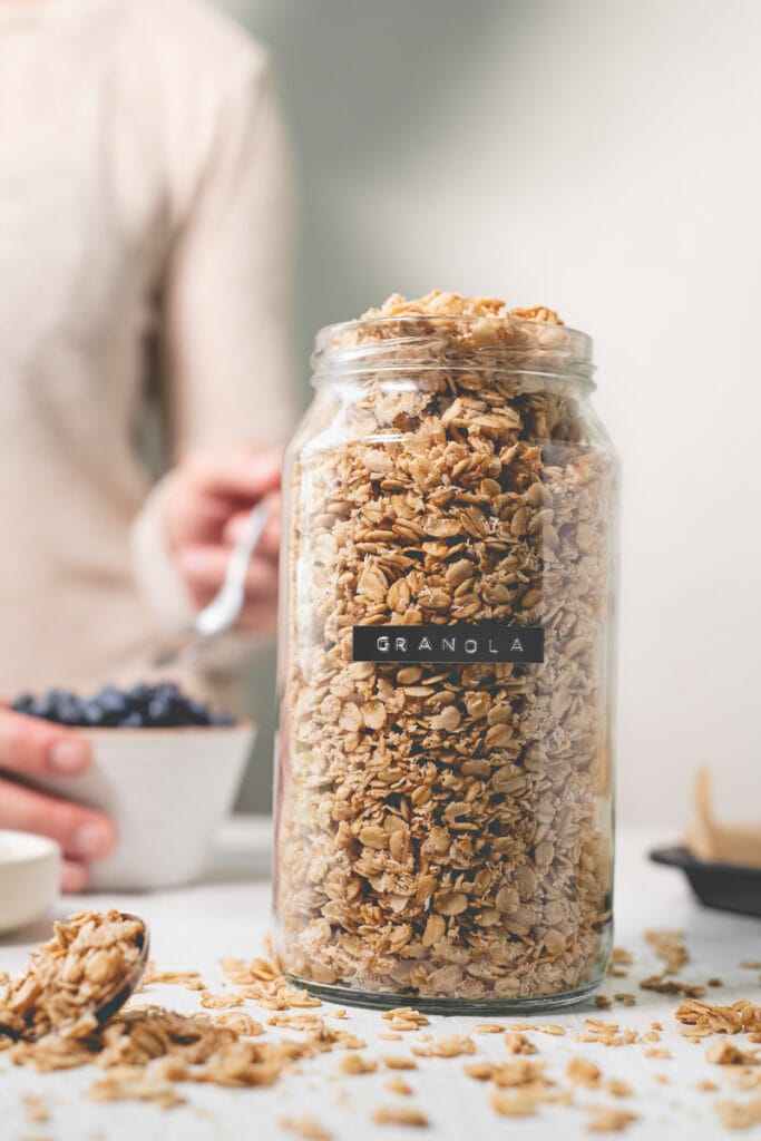 An overflowing of homemade granola sitting next to a spoonful of spilled oats, a bowl of shredded coconut and a heaping bowl of blueberries.