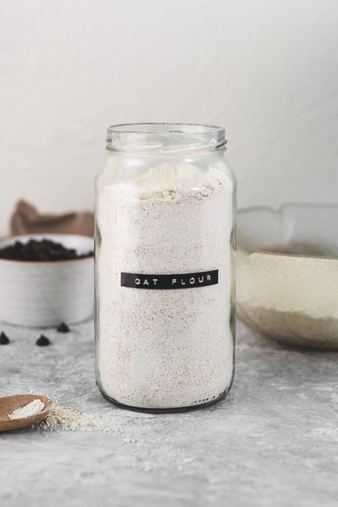 An open jar labeled oat flour sitting on a counter with a mixing bowl, wooden spoon and linen cloth.