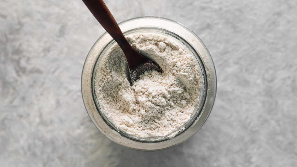 A close-up overhead view of freshly ground oat flour being scooped out of a jar with a spoon.