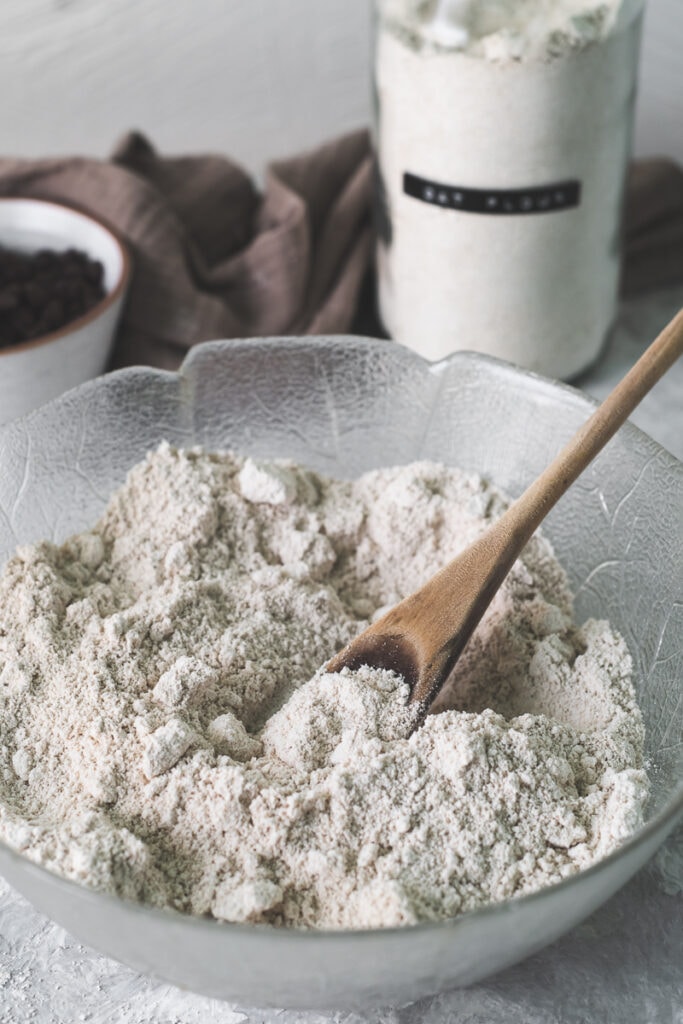 A wooden spoon sunken into  a bowl of fluffy flour, sitting in front of a bowl of chocolate chips, jar flour and a grey-brown linen tea towel.