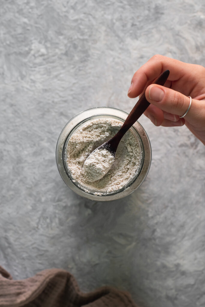 A hand scooping a spoonful of oat flour out of a glass jar.