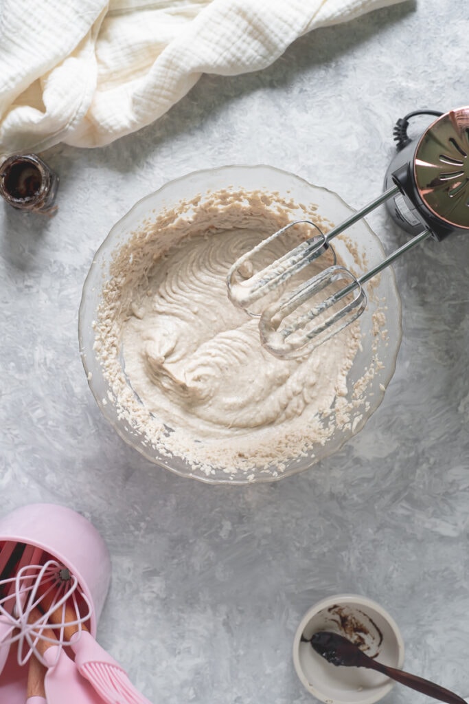 A bowl of whipped cream underneath the dripping beaters of a hand mixer surrounded by vanilla paste, a linen cloth and a pink container full of baking utensils.