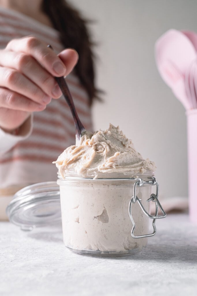 A woman in a pink and white striped shirt dipping a small wooden spoon into a heaping jar of cashew whipped cream and a pink container holding matching utensils in the background.