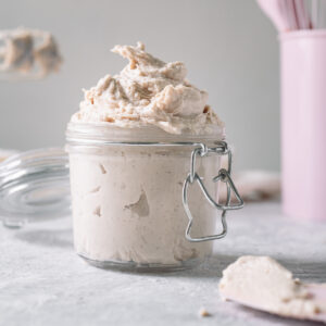 Homemade whipped cream swirled in an open latch jar with freshly used beaters of a hand mixer and a container of pink baking utensils behind.