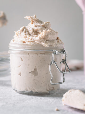 Homemade whipped cream swirled in an open latch jar with freshly used beaters of a hand mixer and a container of pink baking utensils behind.