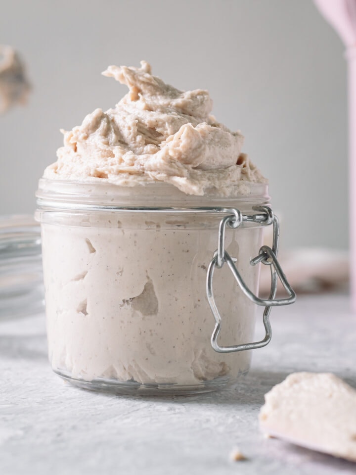 Homemade whipped cream swirled in an open latch jar with freshly used beaters of a hand mixer and a container of pink baking utensils behind.