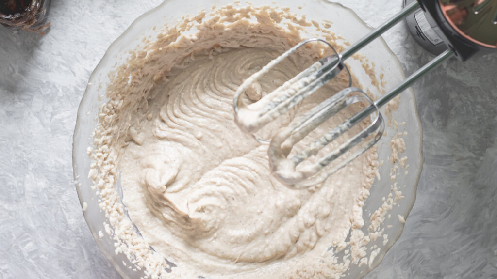 A glass mixing bowl full of freshly whipped cashew cream with the beaters just taken out of the bowl, resting above.
