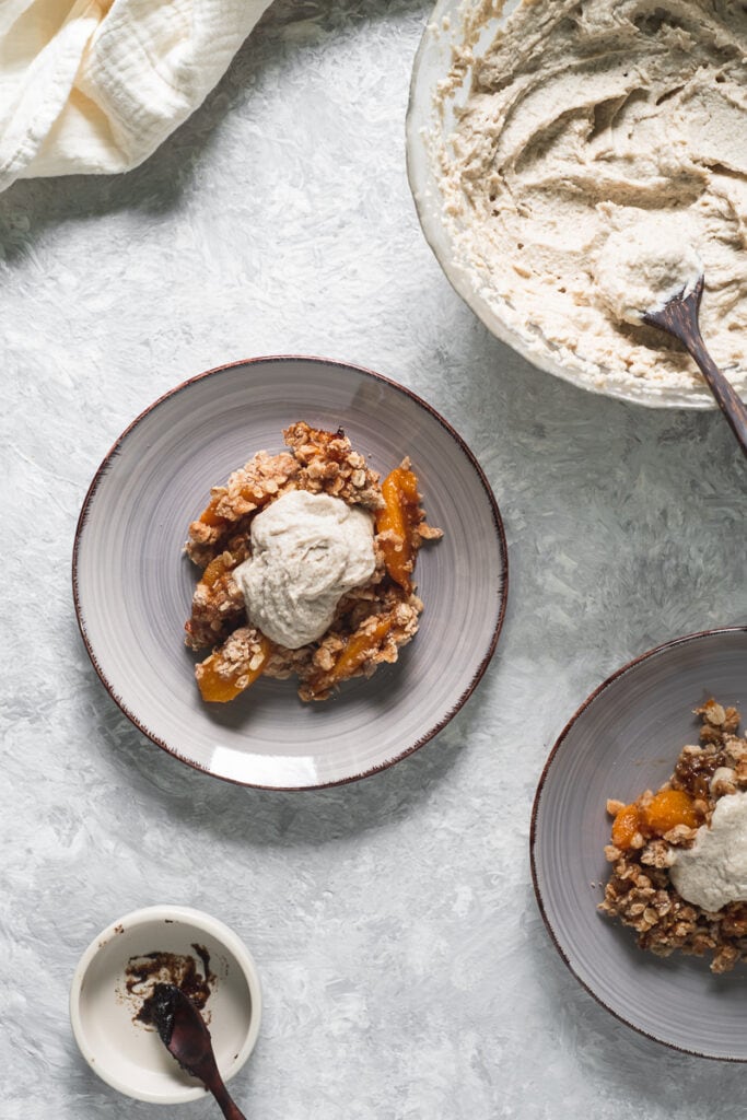 Two plates of peach crumble topped with whipped cream beside a large mixing bowl of more whipped cream, linen cloth and emptied bowl of vanilla paste.