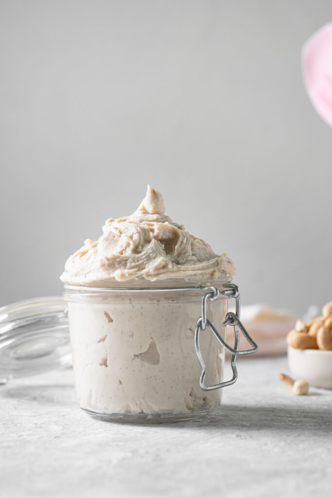 A latch jar overfilled with whipped cream swirled above the rim of the jar and a spilling bowl of cashews behind.