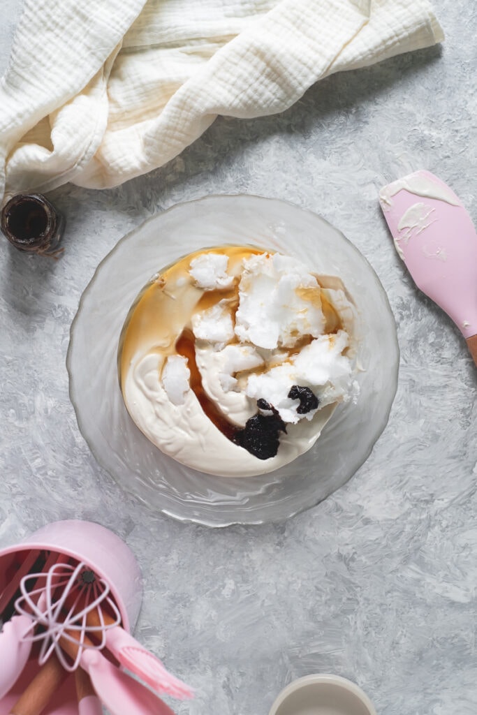 A glass mixing bowl full of cashew cream, maple syrup, coconut oil and vanilla paste surrounded by a pink spatula, linen cloth and jar of vanilla paste.