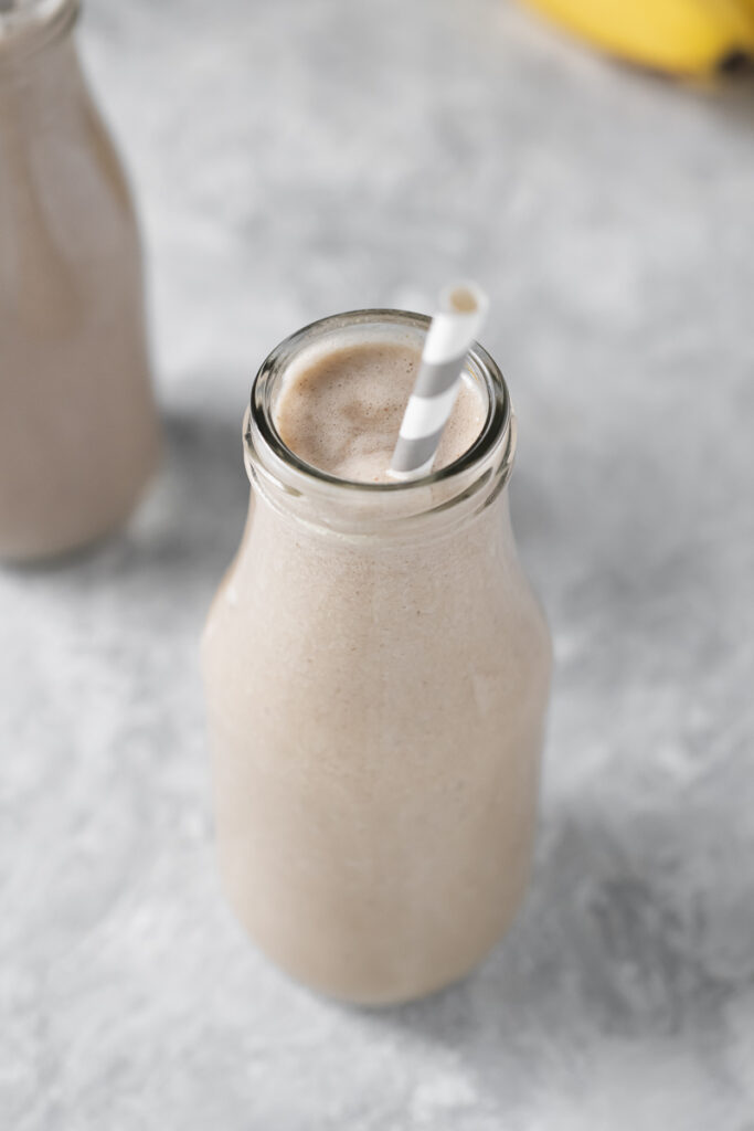 Two vanilla cream coloured smoothies in milk bottle jars with striped straws sitting front of a linen cloth and a bunch of ripe bananas.