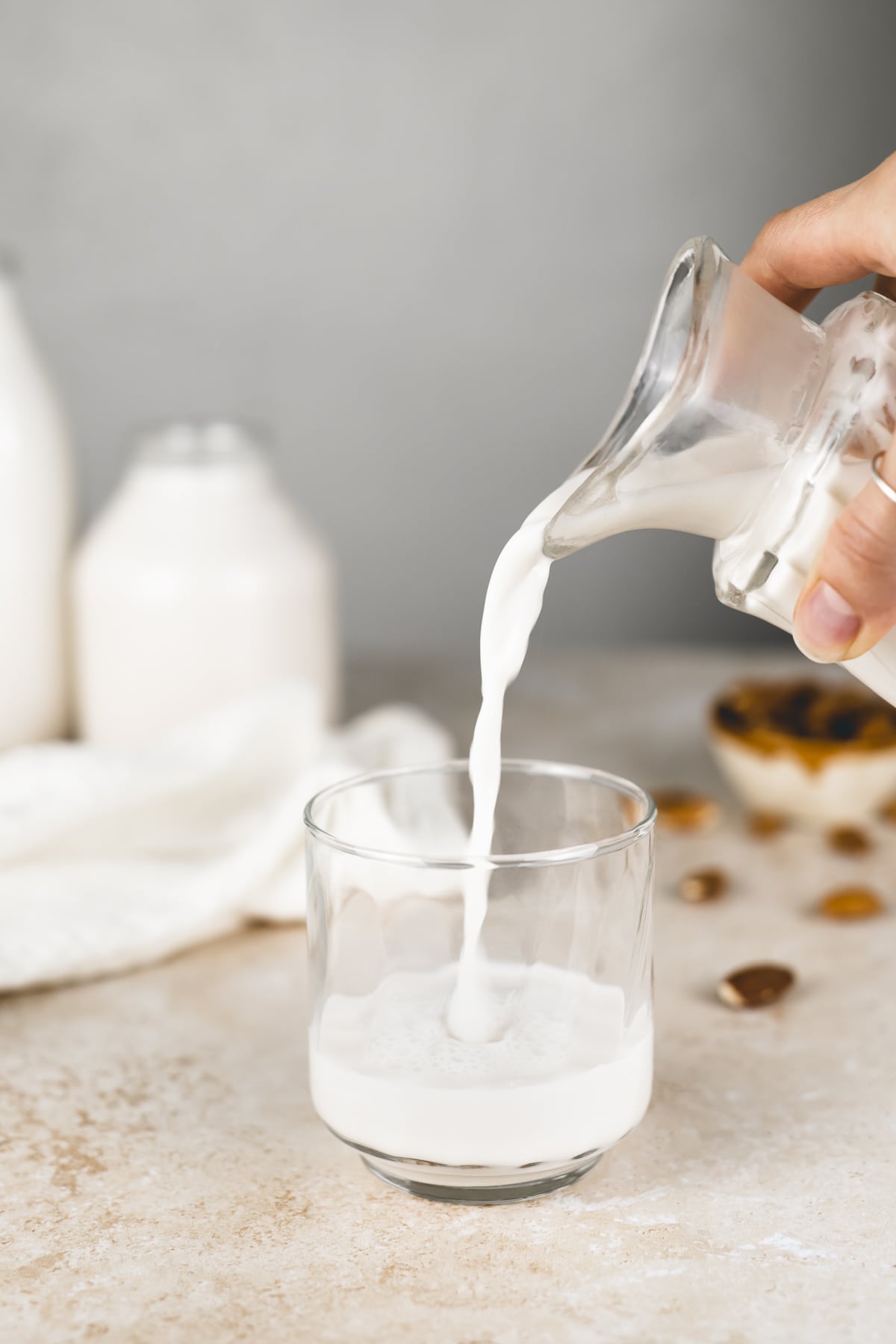 Fresh almond milk being poured from a creamer jug into a glass with an overflowing bowl of almonds and two jugs of milk in the background.