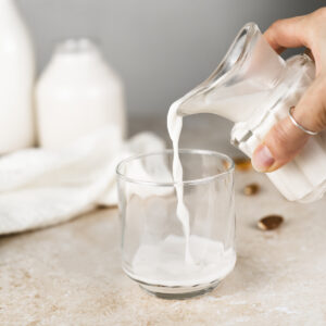 Fresh almond milk being poured from a creamer jug into a glass with an overflowing bowl of almonds and two jugs of milk in the background.