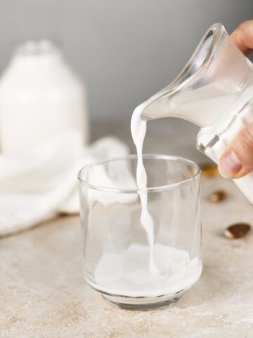 Fresh almond milk being poured from a creamer jug into a glass with an overflowing bowl of almonds and two jugs of milk in the background.
