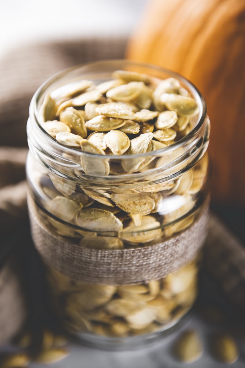 A jar full of roasted pumpkin seeds hugged by a warm coloured linen cloth and orange pumpkin.